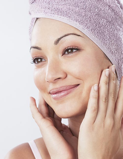 A smiling woman with a towel wrapped around her head touches her cheeks, representing facial aesthetics services in Sandpoint, ID